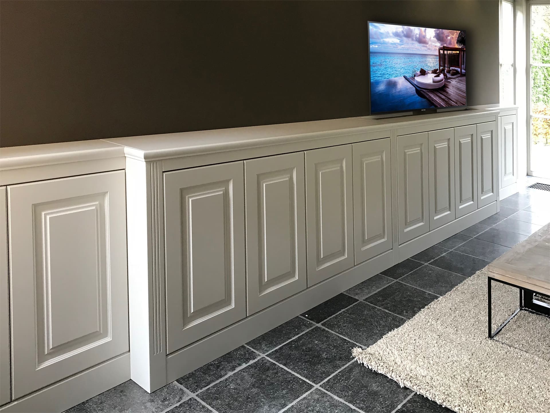 Lange lage witte kastenrij met paneeldeuren onder een donkere wand met televisie, op een blauwstenen vloer