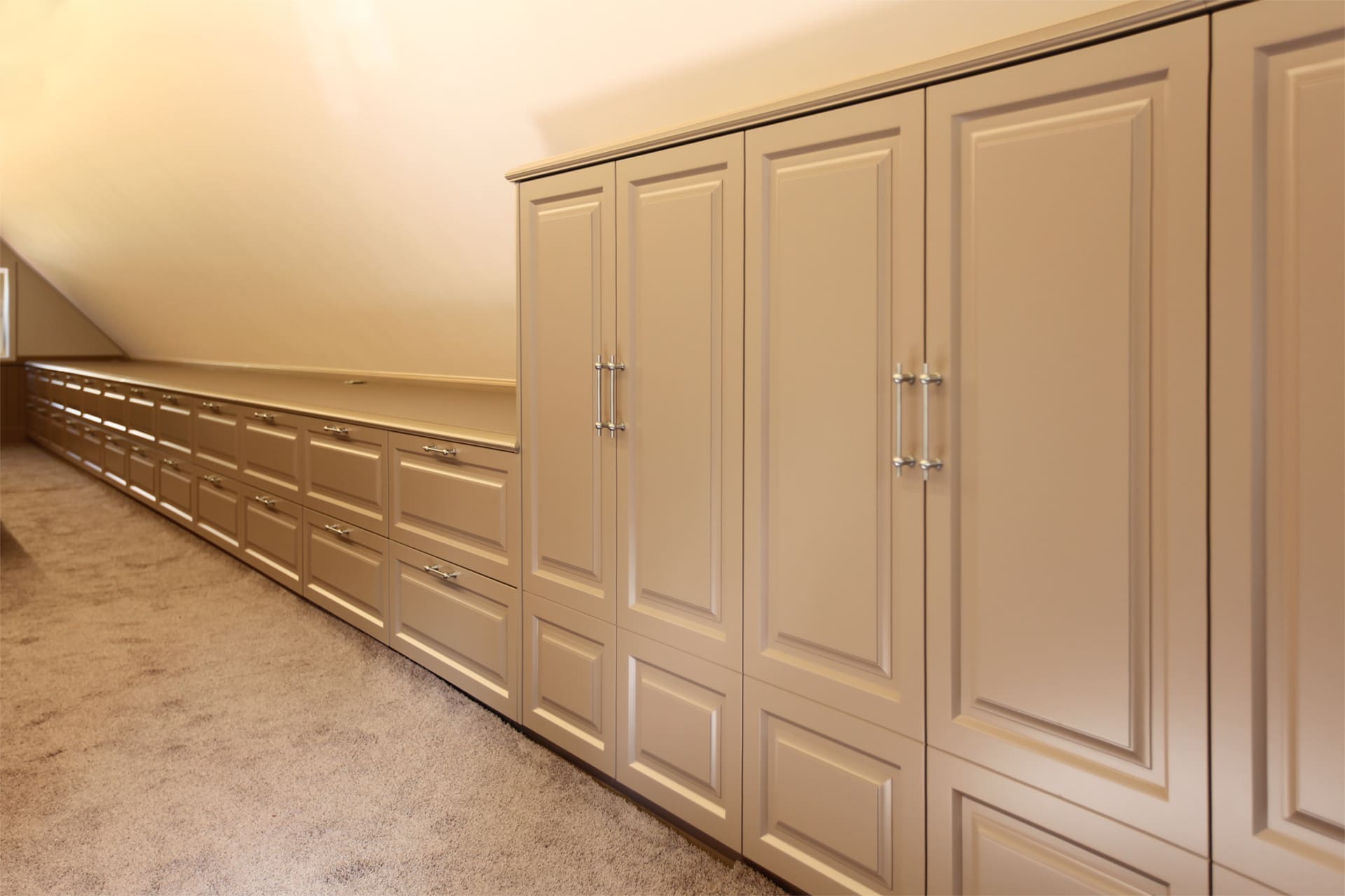 Lange crèmekleurige kastenwand met een doorlopende ladenrij langs een aflopende zolderwand