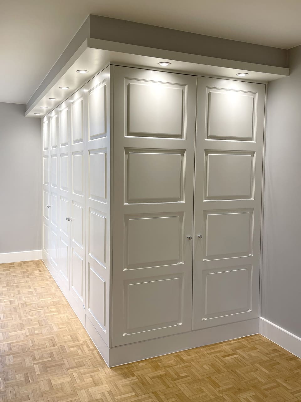 Witte kastenwand met paneeldeuren die in een hoek doorloopt, met een kroonlijst met spots en visgraatparket