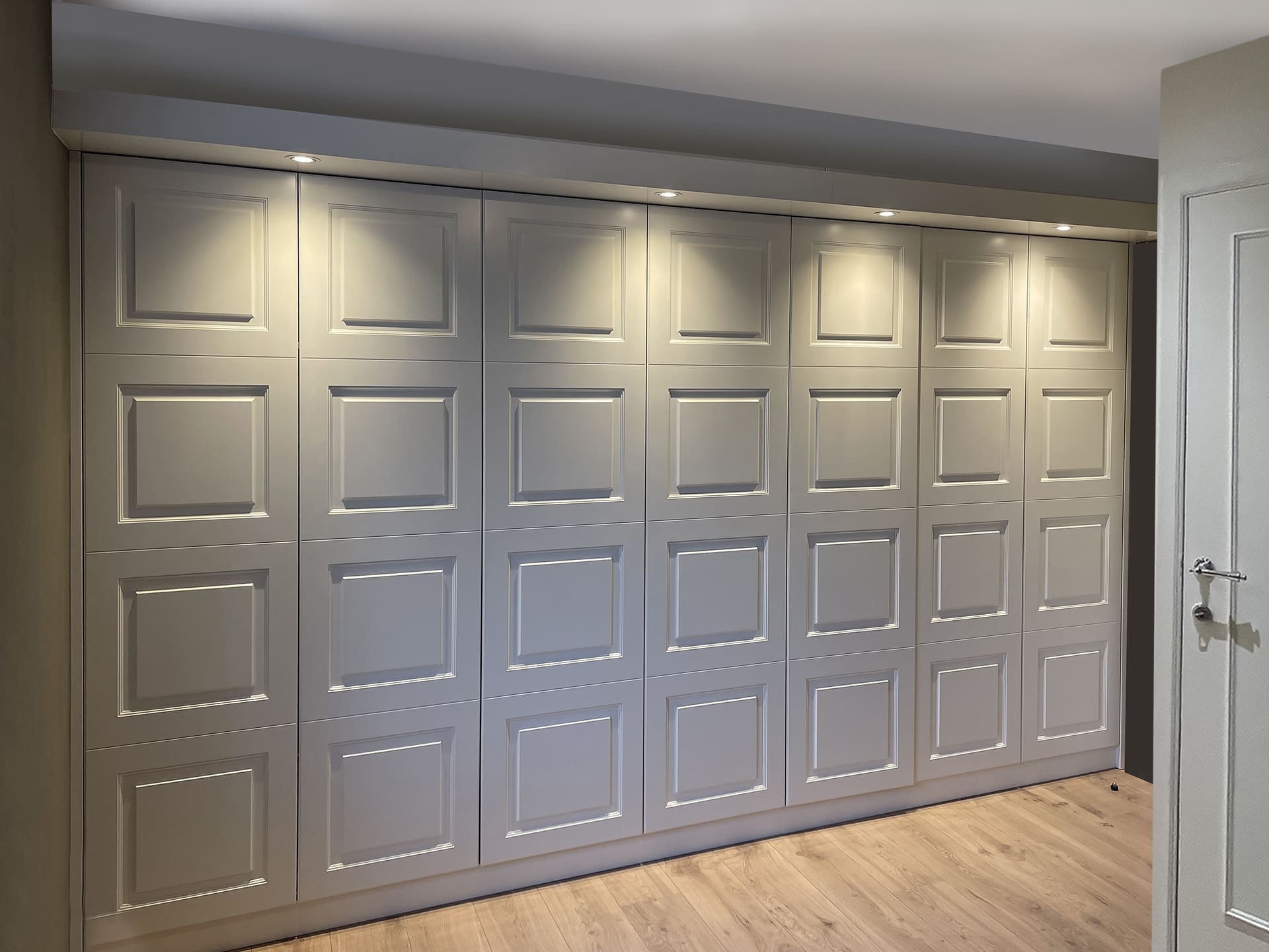 Lange witte kastenwand met paneeldeuren langs een gang, met spots in de kroonlijst