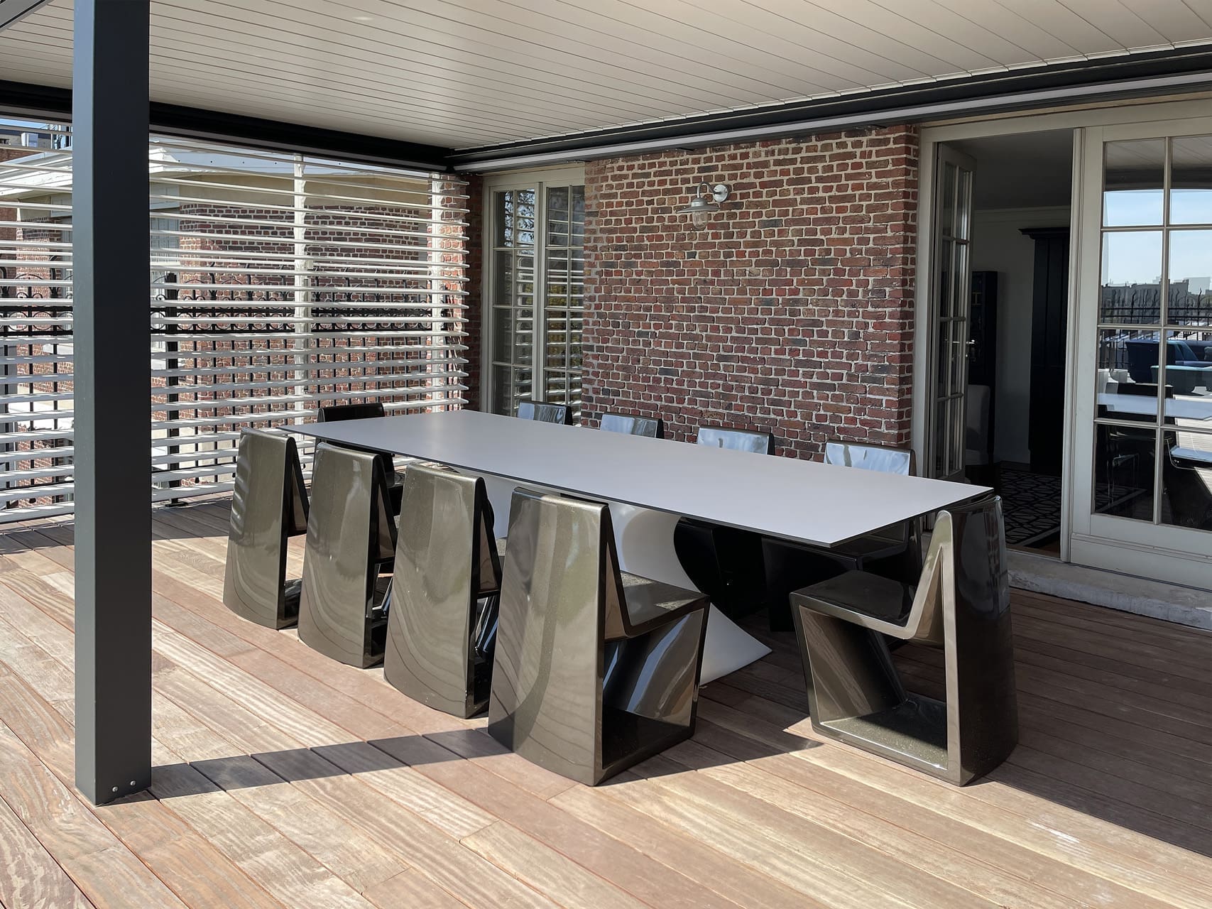 Langer heller Außentisch mit dunklen Designstühlen unter einer Lamellenpergola