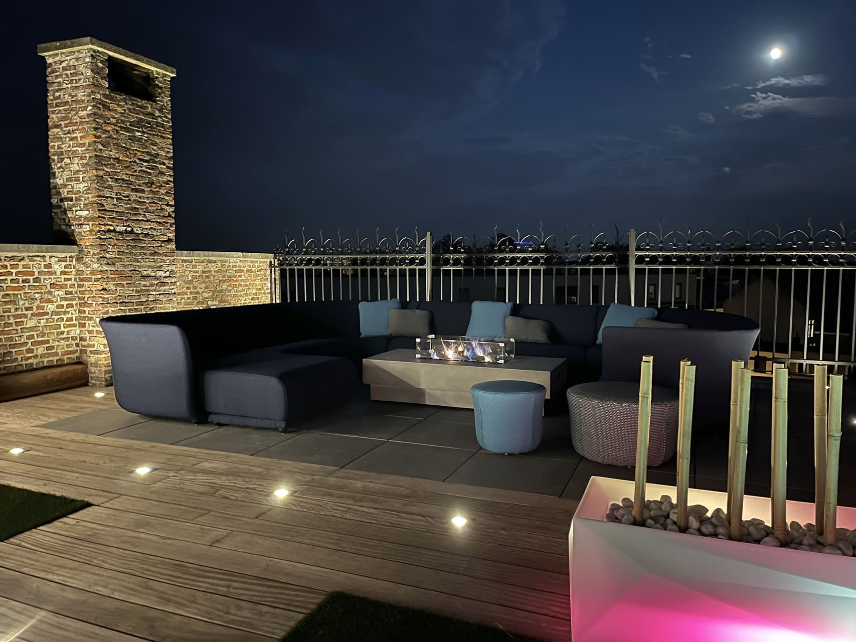 Loungesofa mit Feuertisch auf einer Dachterrasse am Abend, mit einem ausgeleuchteten gemauerten Schornstein