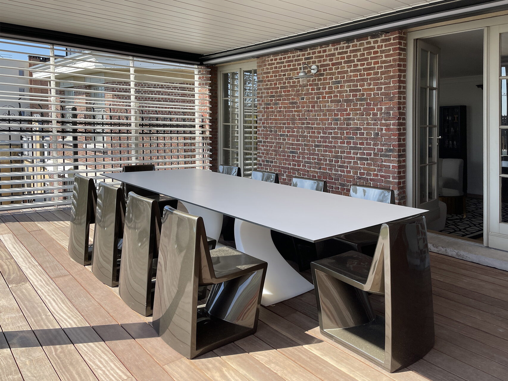 Langer heller Außentisch mit glänzenden Designstühlen auf einem Holzdeck unter einer Pergola