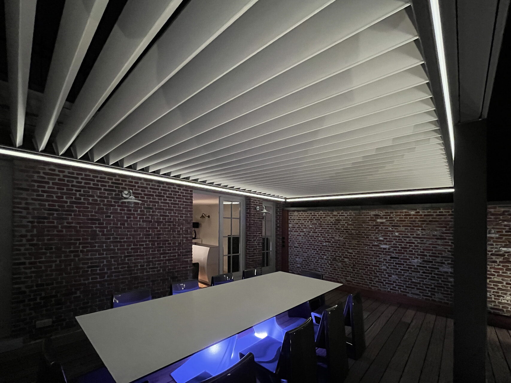 Aluminium-Pergola mit beleuchteten Lamellen über einem langen Außentisch mit blau beleuchteten Stühlen