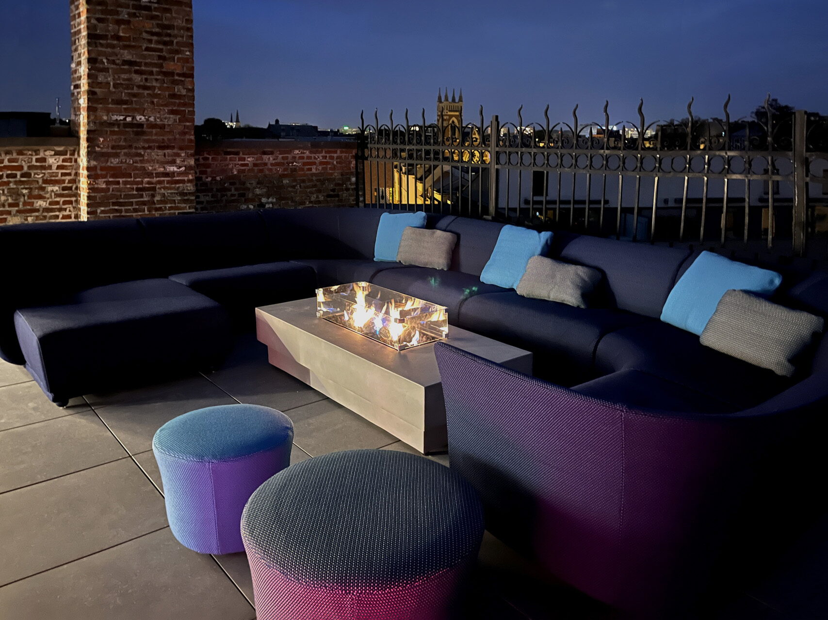 Blaues modulares Outdoor-Loungesofa mit Feuertisch und Poufs auf einer Dachterrasse in der Dämmerung