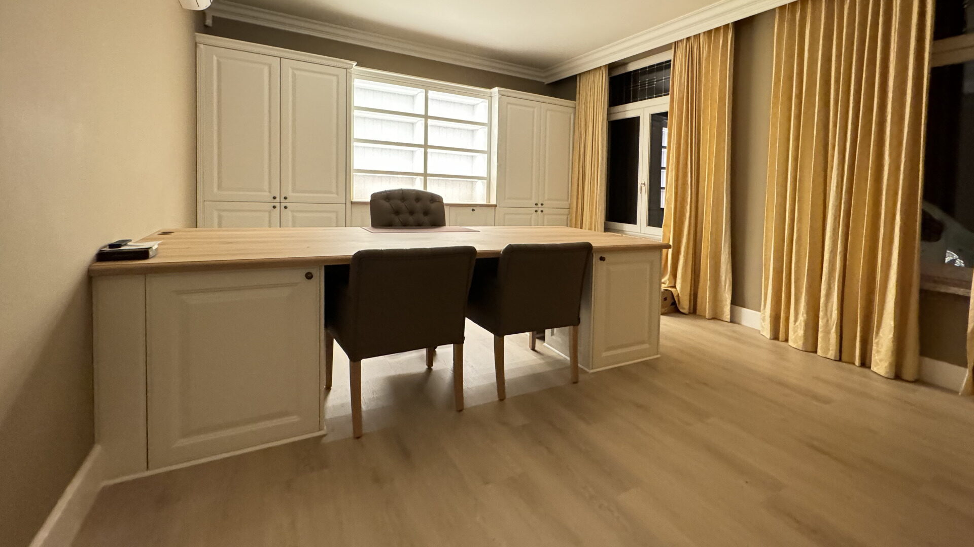 Bureau met een lang eiken blad, crèmekleurige onderkasten, shutters voor het raam en goudkleurige gordijnen