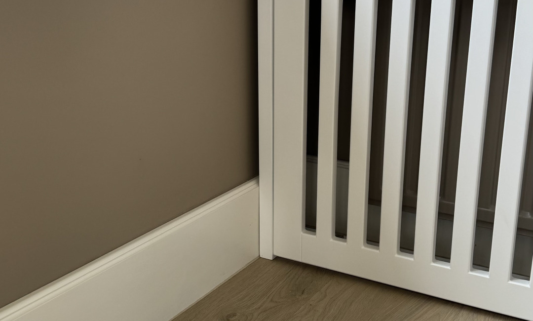 Hoekdetail van een witte radiatorombouw met verticale latten tegen een plint