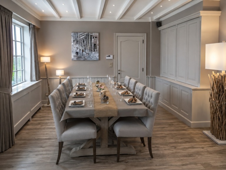 Een lange houten eettafel met tien gecapitonneerde stoelen, met lambrisering langs de wand en een ingebouwde kast tot tegen het plafond.
