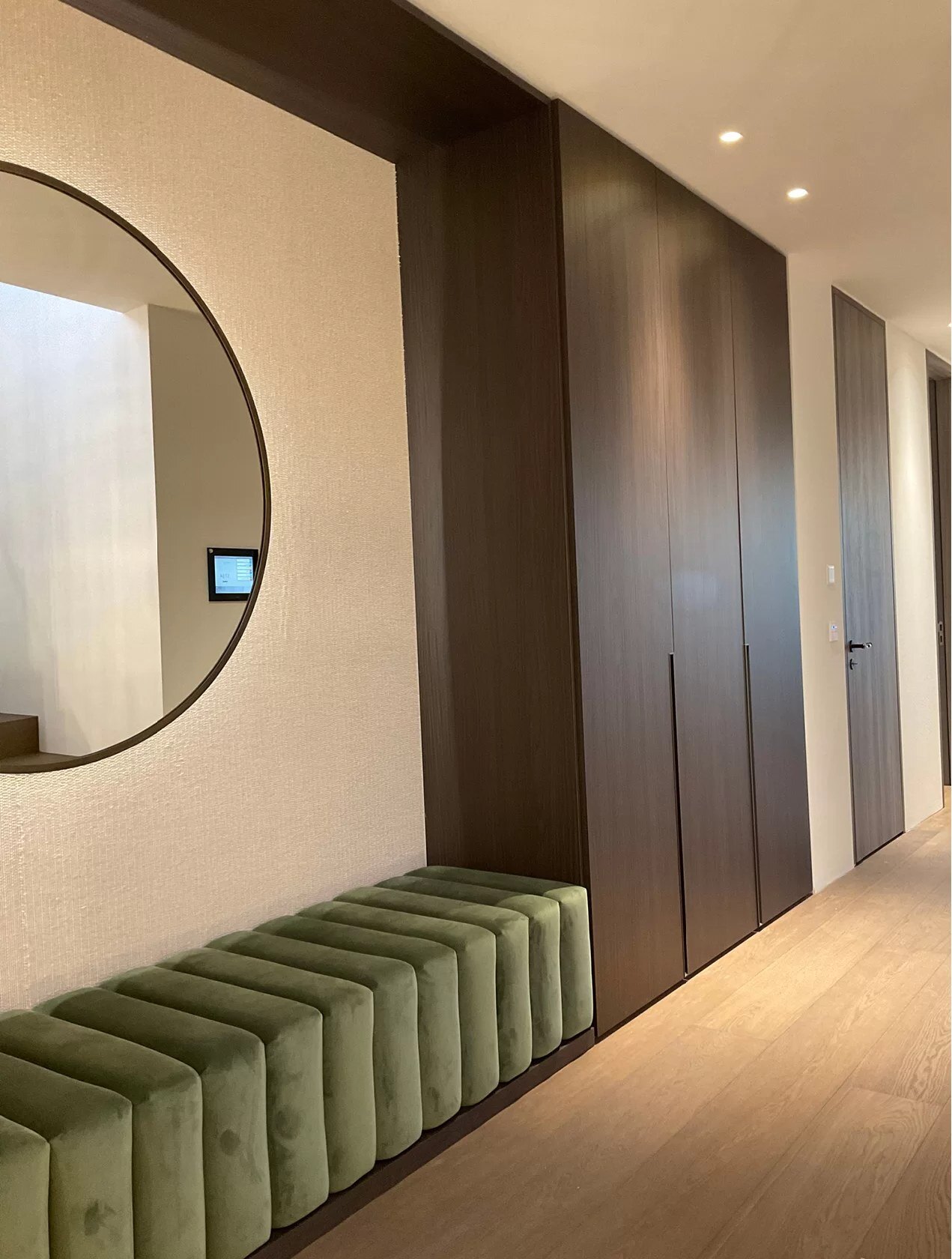 Ronde spiegel op een cremekleurige wand naast een kastenwand in donker hout, met een olijfgroene geribde zitbank