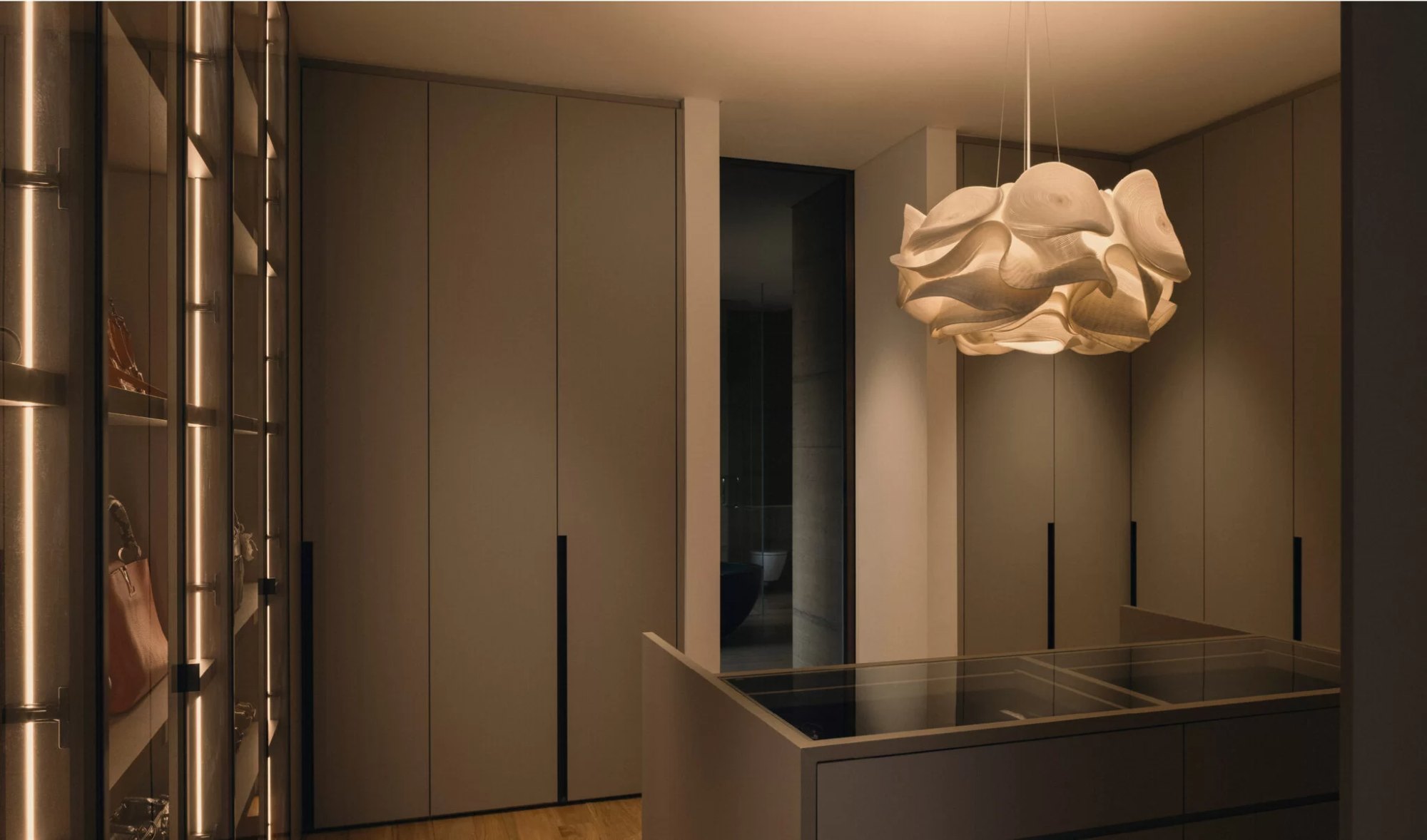 Ein begehbarer Kleiderschrank in warmer Eiche mit Glastüren und einer skulpturalen Hängeleuchte.