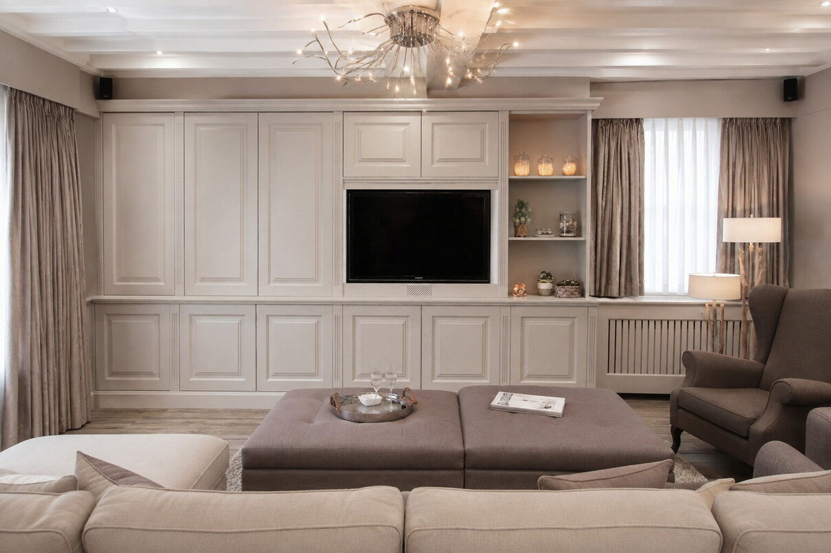 Een wand met gepaneelde kasten van vloer tot plafond, de televisie in een nis in het midden, open legvakken ernaast en een radiatorombouw in dezelfde lak.