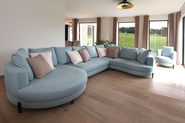 Een grote hoekzetel in lichtblauwe stof met een ronde arm aan één zijde, met een bijpassende draaifauteuil voor de raampartij.