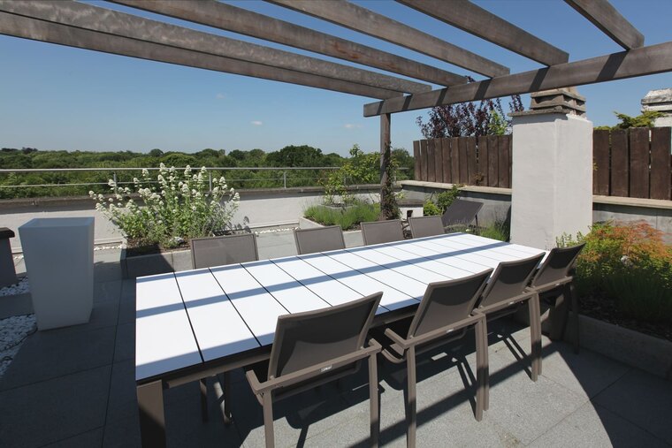 Een dakterras met een pergola boven een lange eettafel met stoelen, met plantenbakken langs de rand.