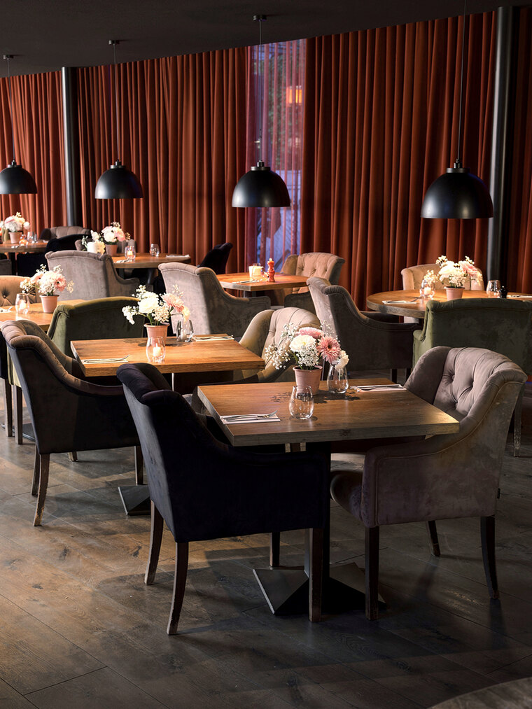 Een restaurantzaal met gestoffeerde stoelen in verschillende tinten rond houten tafels, met zwarte hanglampen laag boven elk tafelblad.