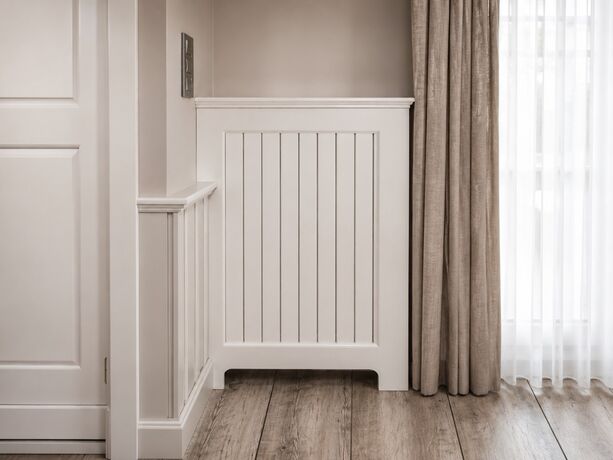 Een radiatorombouw in geschilderd hout met verticale latten, tussen een paneeldeur en een gordijn, op een eiken plankenvloer.