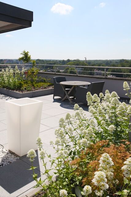 Een dakterras met een verlichte witte kuip op grind, een houten tafel met twee fauteuils, en een brede rand met witte bloemen en heesters.