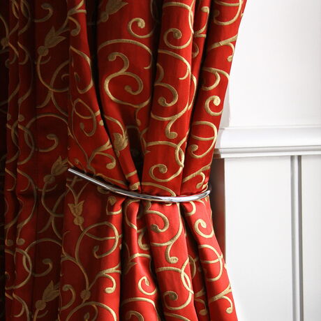 Een rood gordijn met een goudkleurig motief, opzij gehouden door een gebogen metalen haak in de wand.