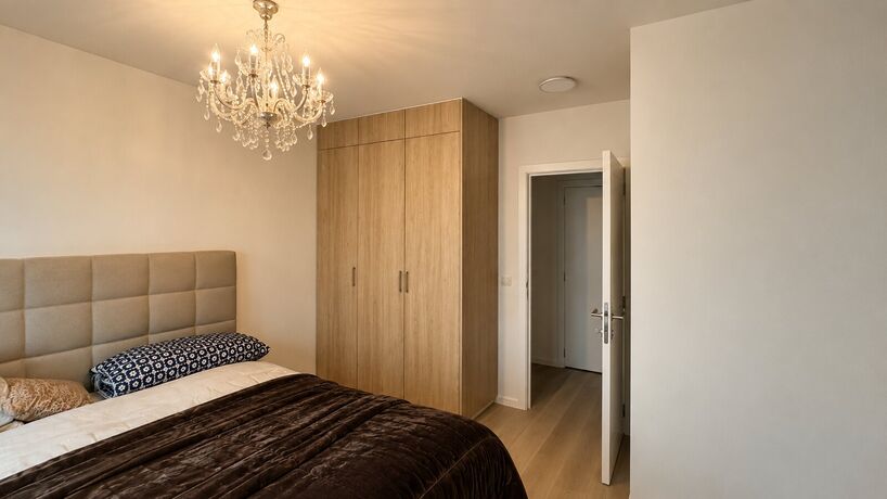 Een kast in eik van vloer tot plafond naast een deur, in een slaapkamer met een gecapitonneerd hoofdbord en een luchter.