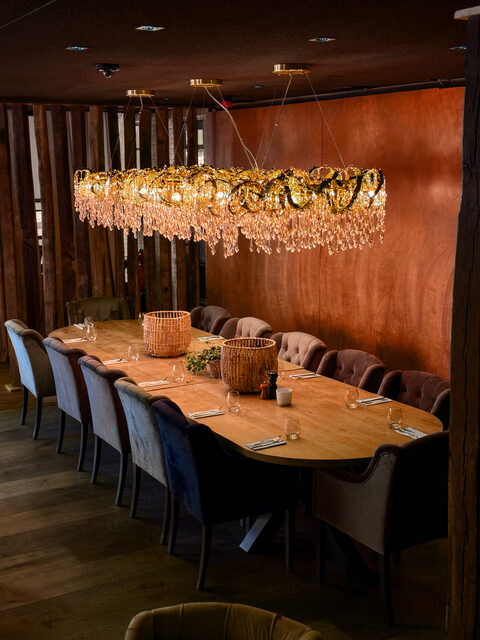 Een lange tafel voor een wand in ruw hout, met een langwerpige luchter in kristal over de volle lengte.