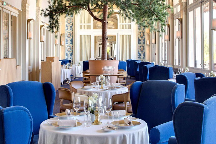 Een restaurantzaal met losse tafels en gestoffeerde stoelen onder een hoog plafond met zuilen.