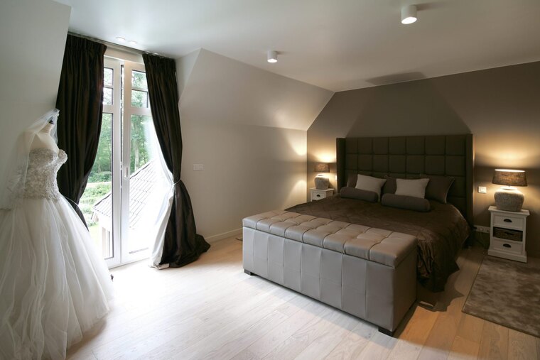 Une chambre sous les toits avec une haute tête de lit capitonnée, une banquette au pied du lit et des rideaux foncés aux fenêtres de toit.