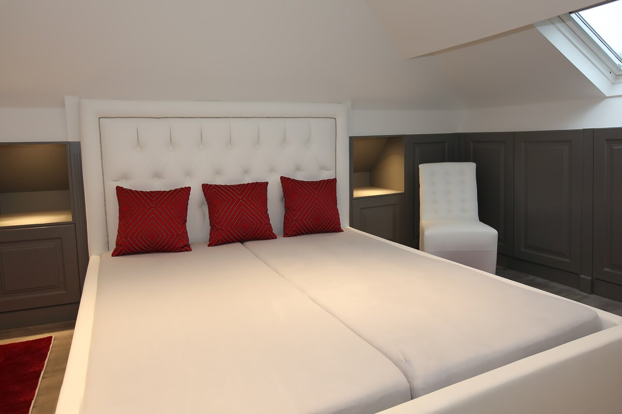Une tête de lit capitonnée blanche avec trois coussins rouges, et à côté du lit une niche éclairée dans le mur d'armoires.