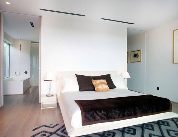 Une chambre claire avec un lit bas, un couvre-lit foncé et une percée vers la salle de bains derrière.