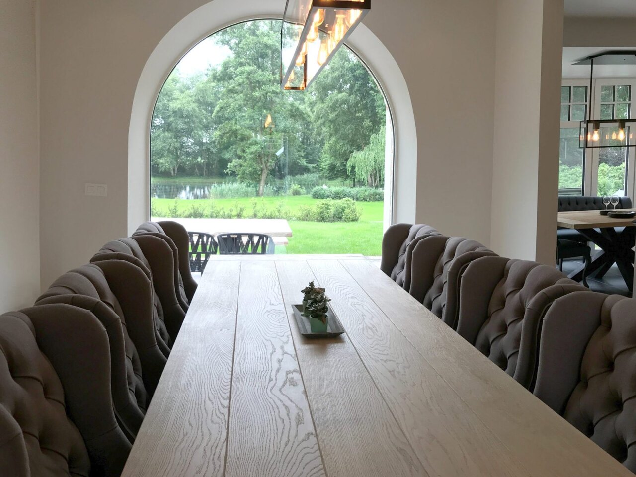 A long oak table with buttoned chairs on either side, with an arched window in the background.