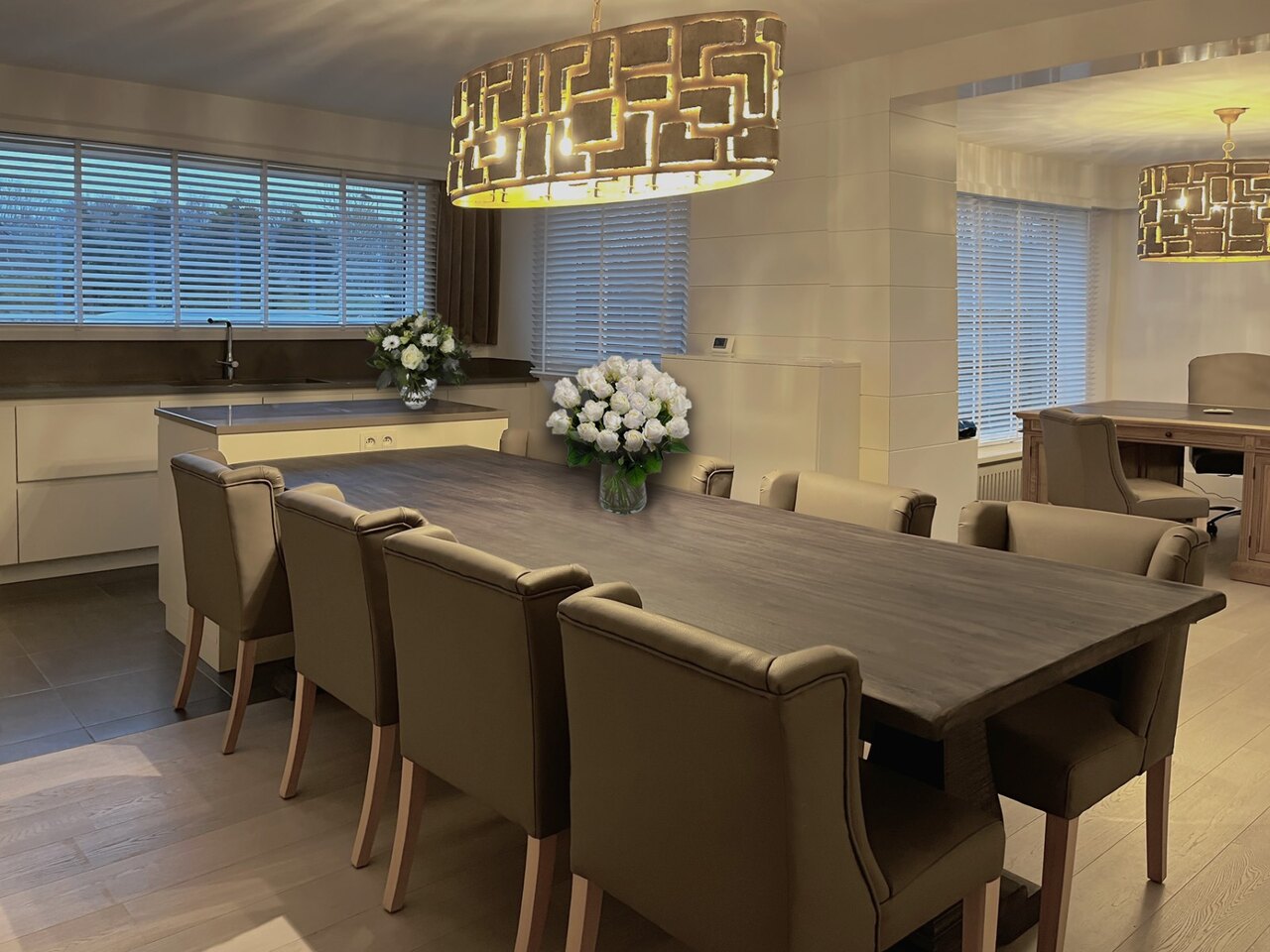 A long table with upholstered chairs connecting onto a kitchen island, with a chandelier above it.