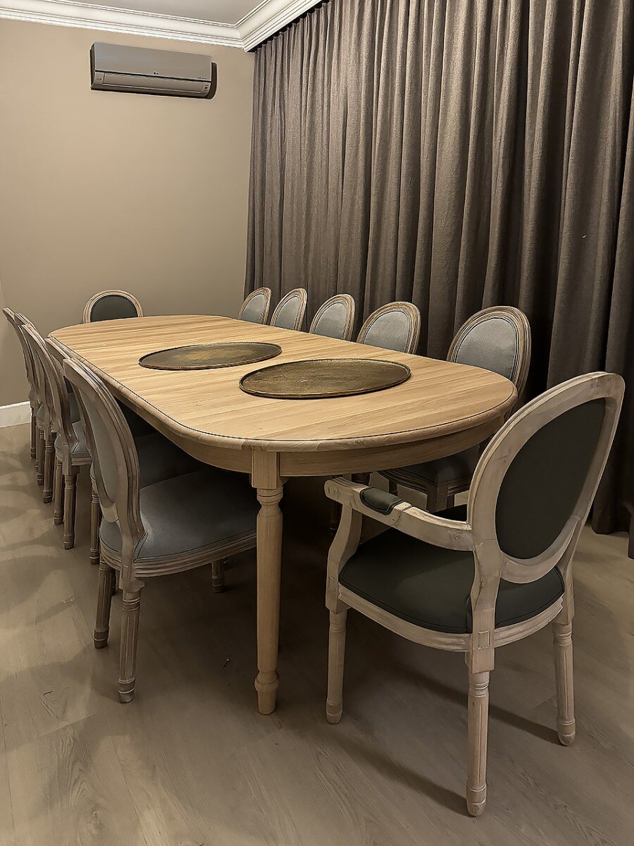 An oval oak table on a central pedestal, with medallion chairs around it and heavy curtains in the background.