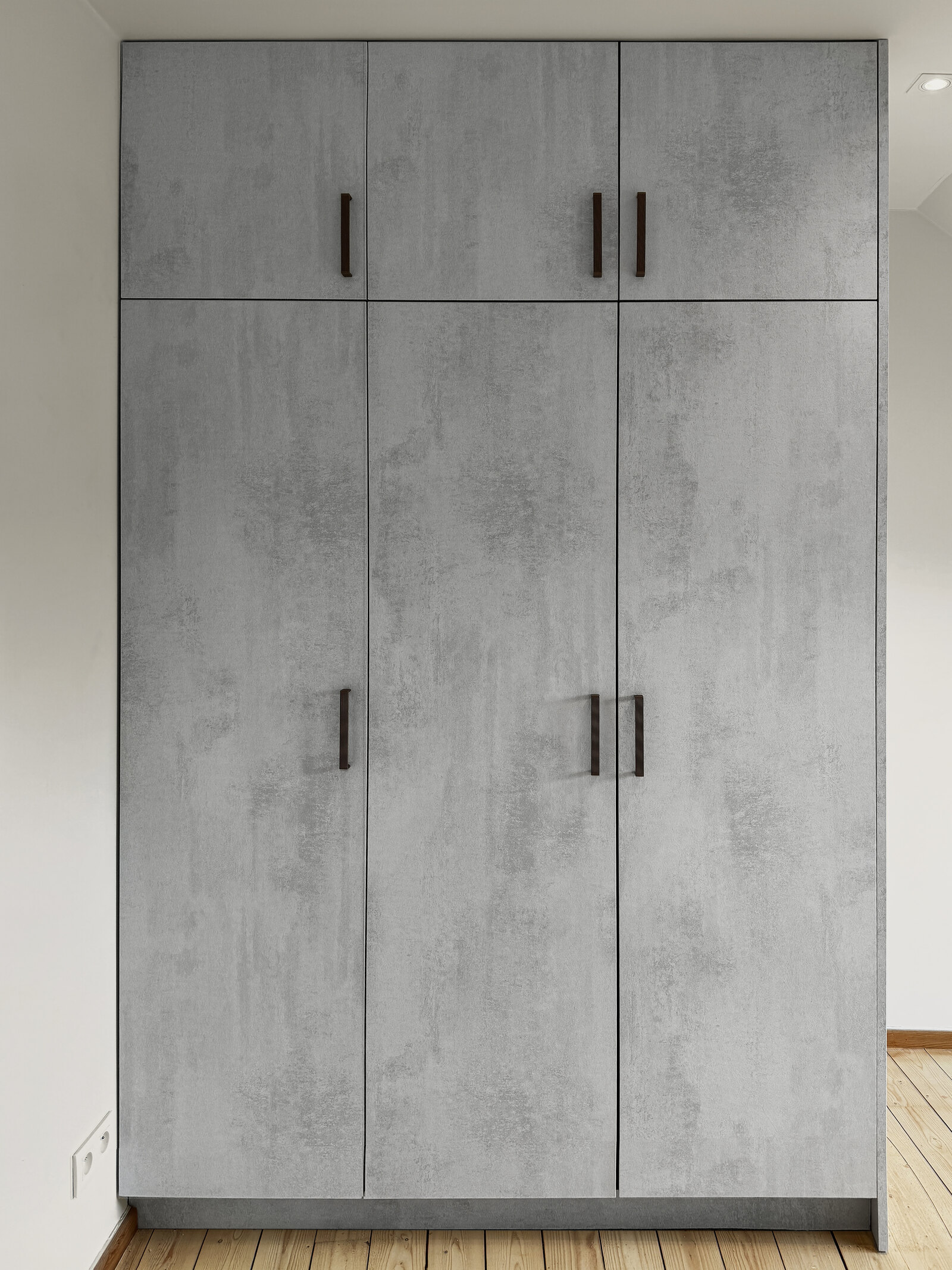The installed concrete-look wardrobe with three doors and bronze handles, in a room with a wooden floor.